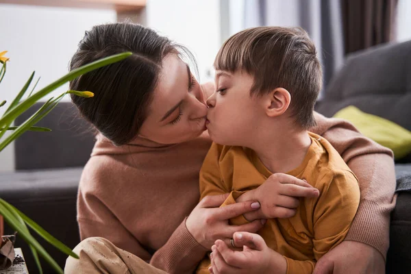Woman kissing at the lips her son with down syndrome while sitting at home at the living room. Family and happy childhood concept