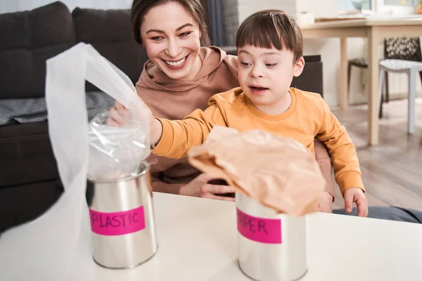 Family of mother and child boy with genetic disorder sorting garbage at home at the table. Concept of recycling. Stock photo