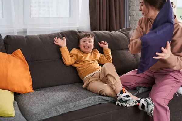 Pillow fight. Overjoyed little boy with genetic disorder laughing out loud while playing with his mother at the living room. Stock photo