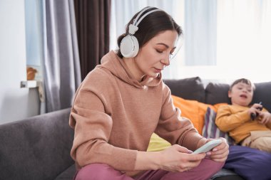 Kanepede sakin bir şekilde otururken akıllı telefon ekranına bakan beyaz bir kadın. Arka plandaki çocuğu. Mutlu aile kavramı. Stok fotoğrafı