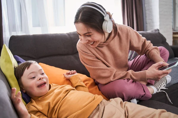 Cheerful woman holding smartphone and laughing out loud to her son with genetic disorder while relaxing at the couch together