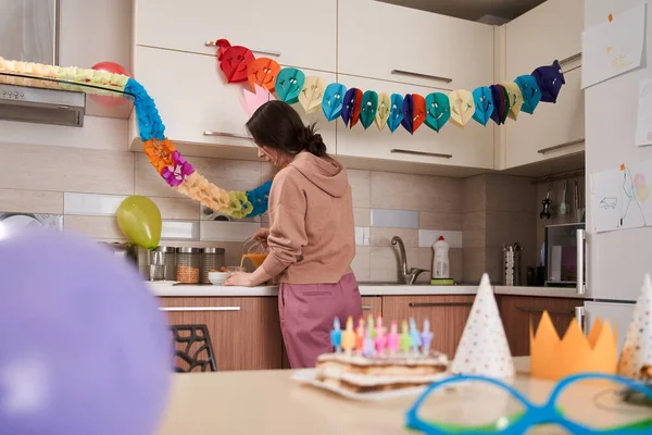 Birthday cake with candles standing at the table with another party decorations nearby. Mother standing at the kitchen at the background