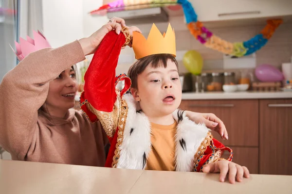 Little boy with genetic disorder wearing king costume preparing to the party with his mother while sitting at the table at the kitchen. Family concept