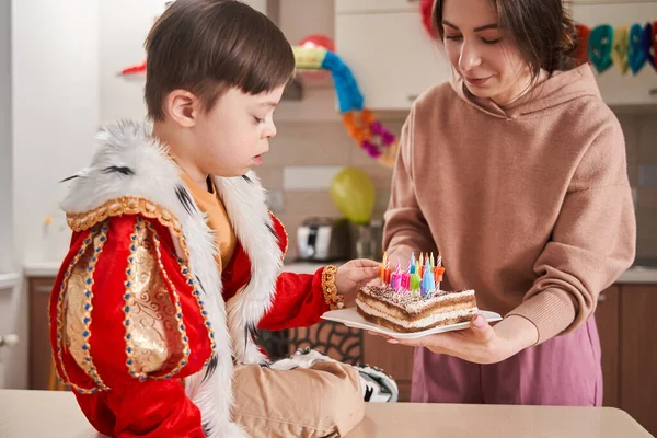Little boy with genetic disorder sitting at the table and examining his birthday cake with candles while preparing to his anniversary party