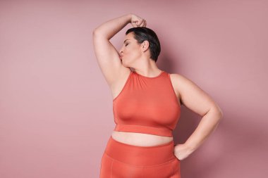 Pembe arka plandaki eğitimden sonra genç beyaz tenli ve iri yarı bir kadın modelin öpüşme kasları. Spor anlayışı, sağlıklı yaşam tarzı, vücut pozitifliği, eşitlik