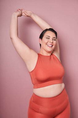 Genç, pozitif, gülümseyen, şişman kadın spor kıyafetleri, pazılarını gösteriyor, pembe stüdyo geçmişine karşı egzersiz yapıyor. Spor anlayışı, vücut pozitifliği, kilo kaybı, vücut ve sağlık hizmetleri