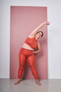 Stüdyo pembe arka planda izole edilmiş spor kıyafetleri giymiş şişman, güler yüzlü bir kadının egzersiz yaptığı uzun bir fotoğraf. Spor, spor ve vücut pozitifliği
