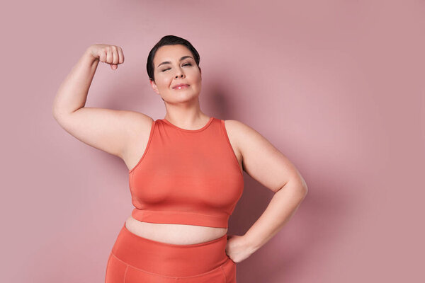 Cheerful large woman showing biceps on pink background. Plus size female in sports clothing looking at camera and smiling