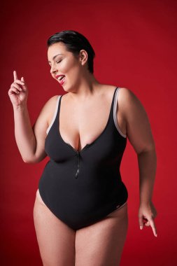 Ara veriyoruz. Mutlu büyük beden kadın siyah iç çamaşırı giyiyor, gözleri kapalı dans ediyor, hareket ediyor ve müzik dinliyor, kırmızı stüdyo arka planında izole edilmiş bir şekilde rahatlıyor.