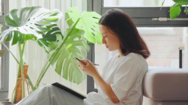 Kanepede otururken not alan esmer kızın yakın görüntüsü. Asyalı kız evde ders çalışırken akıllı telefona bakıyor.