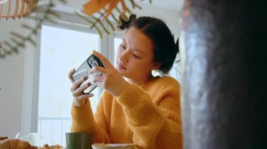 Konsantre Asyalı kız mutfakta akıllı telefonuyla ilginç videolar izlerken yemek yiyor. İnsanlar ve teknoloji kavramı