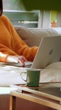 Bilinmeyen bir kadının dizüstü bilgisayar yazarken ve oturma odasındaki kanepede otururken çalışırken dikey görüntüsü. Ev hayatı konsepti