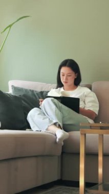 Elinde not defteriyle kanepede oturan ve uzaktan tek başına çalışırken plan yazan Asyalı genç kadının dikey görüntüsü