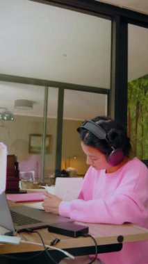 Asyalı liseli kız öğrencinin dikey görüntüsü kulaklık takmış internetteki Webinar dersini izliyor odaklanmış suratlı dizüstü bilgisayara bakıyor. Genç kadın videosu öğretmeni uzaktan arıyor.