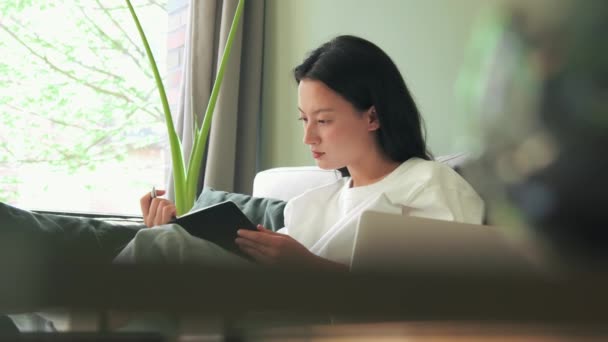 Skupiony Azji Młoda Kobieta Siedzi Kanapie Notatnikiem Rękach Pisanie Planu — Wideo stockowe