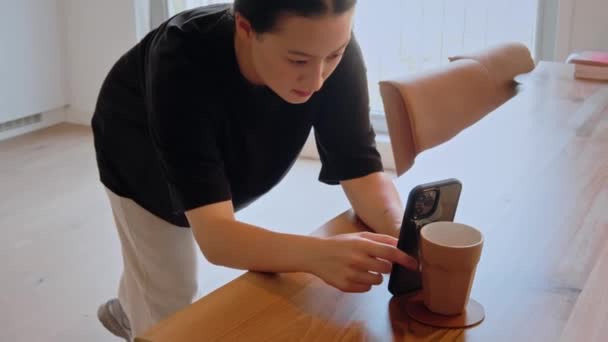 テーブルの上にスマートフォンを持っているかわいいアジアの女の子と光居心地の良いキッチンに立っている間の設定を修正します ブルネット女性一人で時間を過ごす — ストック動画