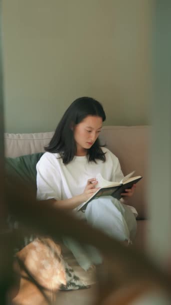 自信に満ちたフレンドリーな若い女性の垂直ビュー彼女の手でノートとソファに座ってリモートで勉強しながらノートを作る — ストック動画
