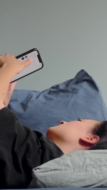 アパートでリラックスしながら携帯電話の画面を見てベッドに横たわっているアジアの女性の垂直方向のビュー テクノロジーの概念 — ストック動画