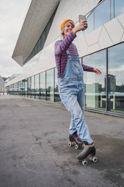 Şehir merkezinde paten kayarken cep telefonuyla selfie çeken aşırı mutlu kadının dikey görüntüsü. Hobi konsepti