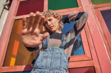 Sarışın, kıvırcık saçlı, şık elbiseli, el kol hareketi yapan ve güneşli caddede kameraya bakan adam. Sokak modası konsepti