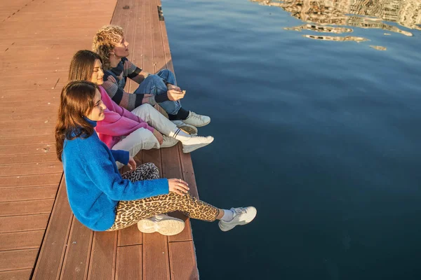 木製の桟橋に座って水の景色を楽しみながら話している幸せな友人たち 若者のライフスタイルのコンセプト — ストック写真