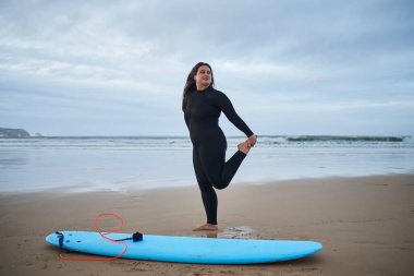 Esnek, genç, beyaz bir kadın deniz kıyısına uzanıyor ve sörf tahtasının yanında gülümsüyor. Spor ve insanlar konsepti