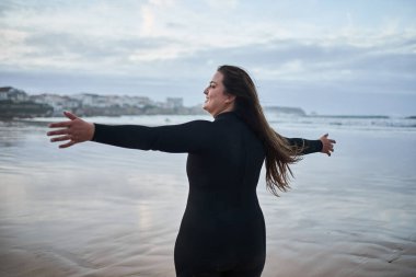 Dalgıç kıyafetli mutlu vücut pozitifliği olan kadın kollarını açıyor ve sörf yaptıktan sonra okyanus sahilinin tadını çıkarıyor. Spor ve eğlence konsepti