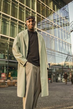 Tek başına yürürken sokak mimarisinde poz veren modaya uygun giyinmiş çok kültürlü bir adamın tam boy görüntüsü.