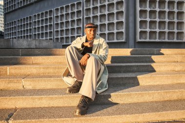 Çok kültürlü, modaya uygun güneş gözlüklü bir adam bahar caddesinde poz verirken merdivenlerde oturuyor. Şehir moda kavramı