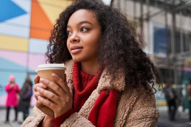 Moda kıyafetleri içinde gülümseyen genç bir kadın sıcak bir içki tutuyor ve mesafeyi arıyor. Sokakta yürüyor ve sıcak içeceğin tadını çıkarıyor.