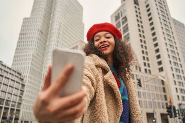 Pozitif kadının arka planda gökdelenlerle sokakta yürürken cep telefonunda selfie çektiği düşük açı.