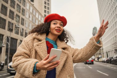 Güzel, çok kültürlü bir kadın sokakta yüksek binalarla taksi tutarken elini kaldırıyor. İnsanlar ve ulaşım kavramı