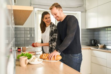 Güzel, mutlu, genç bir çift evdeki mutfakta yemek pişiriyor. Ekmek kesen adam ve karısı ona gülümseyerek bakıyor.