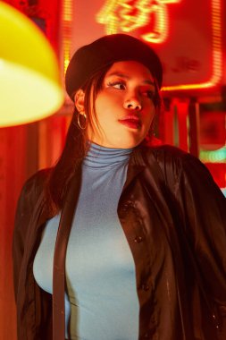 Şehirdeki gece hayatının tadını çıkaran, modaya uygun giyinmiş, mesafeli Asyalı bir kadının dikey görüntüsü. Neon şehir aydınlatmasıyla sokakta duran muhteşem moda hipster kızı.