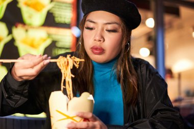 Asyalı bir kadın akşam sokak kafesinde çubuklarla erişte yiyor. Odaklanmış sakin bayan, akşam yemeğinin tadını çıkarıyor.