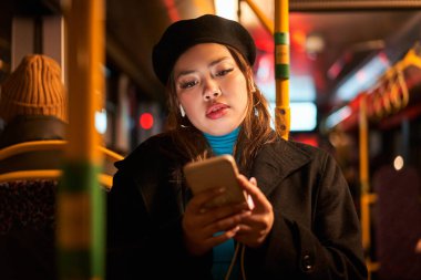 Çekici Asyalı kadın gece otobüse binerken akıllı telefon kullanıyor. Genç ve güzel bir kadın toplu taşıma araçlarını kullanıyor. Şehir ışıklarıyla aydınlatılmış