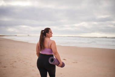 Okyanus sahilinde yoga minderiyle durup suya bakarken gülümseyen pozitif kadın. Stok fotoğrafı