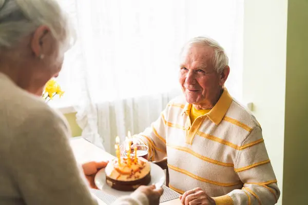 The wife brought a cake with candles to her elderly husband, and the birthday celebrant is ready to make wishes and blow out the candles.