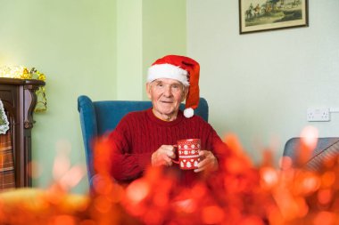 Gülümseyen, sandalyeye oturmuş, arka planda Noel dekoruyla kameraya bakan mutlu yaşlı bir vatandaşın portresi..