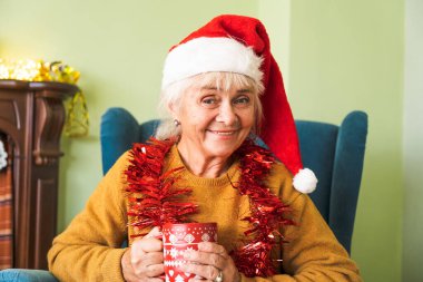 Gülümseyen yaşlı bir kadının portresi, bir sandalyede oturuyor, arka planda bir Noel dekoru ile kameraya bakıyor..