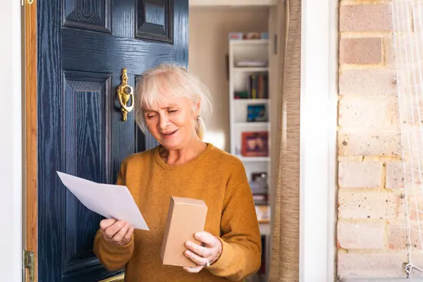 An elderly woman retrieves a delivery from her doorstep, consisting of a small box and an envelope with a letter.