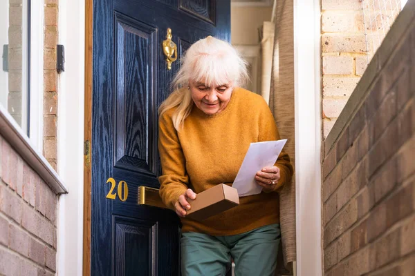 An elderly woman retrieves a delivery from her doorstep, consisting of a small box and an envelope with a letter.