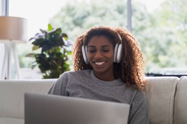 Çekici Afrikalı Amerikalı kadın oturma odasında bilgisayar kullanıyor ve kulaklıkla müzik dinliyor. Teknoloji ve insanlar kavramı