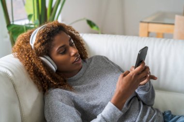 Esmer, çok ırklı kadın. Günlük ev kıyafetleri giyiyor. Kablosuz kulaklıkla koltukta dinleniyor. İnsanlar ve teknoloji kavramı
