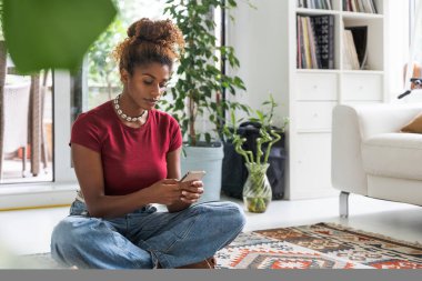 Serene sakin genç kadın halıda dinleniyor ve akıllı telefon uygulamasında internette geziniyor. Halk yaşam tarzı konsepti