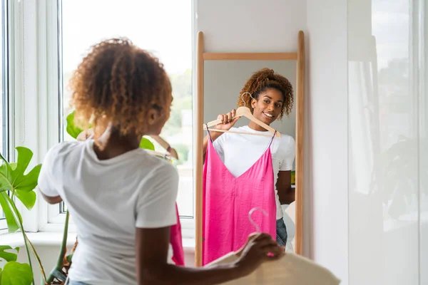 Smiling curly woman trying on clothes and looking at her reflection at mirror. People lifestyle concept