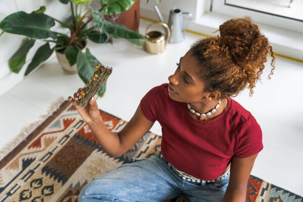 Милосердная молодая женщина, держащая Санту Пало Вуд и медитирующая в уютной комнате. Она сидит в позе лотоса. Полная релаксация