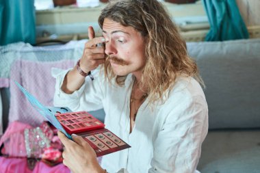 Uzun saçlı adam evde oturuyor ve gözleri tutuyor. Sadece parmaklarıyla makyaj yaparken paleti gölgeliyor. İnsanlar görünüm kavramı