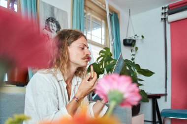Hazırlanırken uzun saçlı, bıyıklı, ruj süren ve dudak boyayan genç bir adam. Aynaya bakan yakışıklı adam.