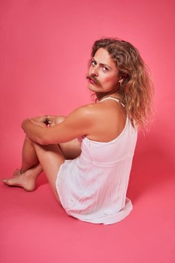 Ciddi, uzun saçlı, bıyıklı, omzuna bakan bir adam stüdyoda pembe arka planda poz verirken.